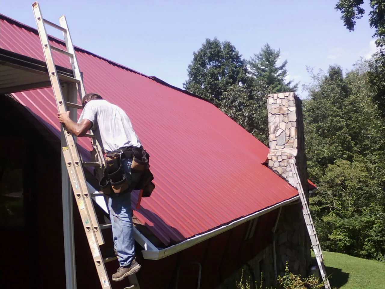 Roofer on a ladder beside a red metal roof and stone chimney — Soffit & Fascia Repair by Roofing Brothers in Bladensburg